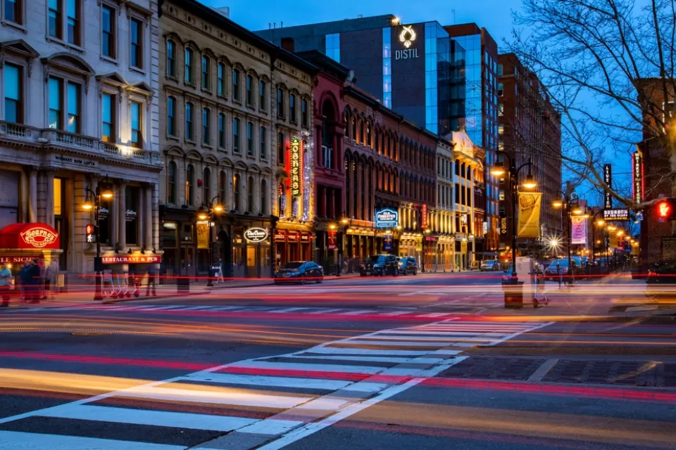 A Downtown Louisville street at night, with the lights of passing shown with a blurred effect.